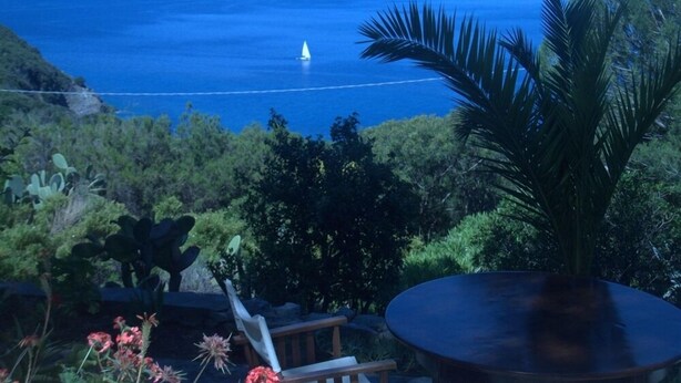 A coastal view with a sailboat on the water, surrounded by lush greenery and a wooden deck with a round table. - House With Sea View On Elba Island - 10 Min Walk from Beach (La Guardia)