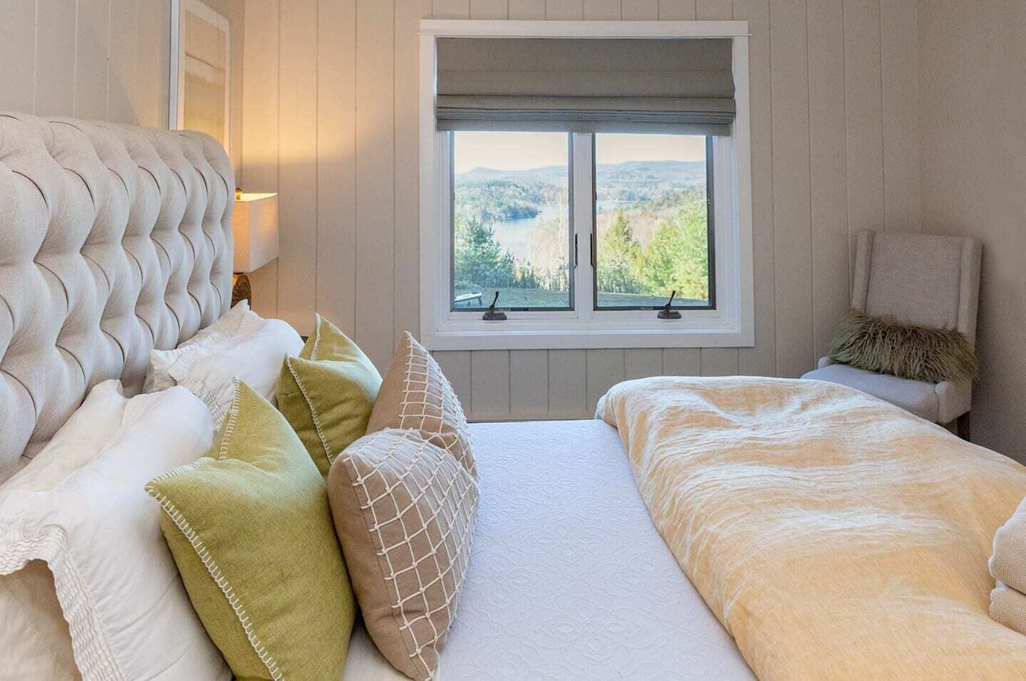 A bedroom with a tufted headboard, a large window with a view of greenery, and a bed with a white and beige duvet.