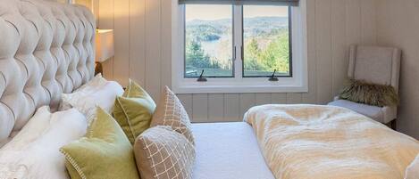 A bedroom with a tufted headboard, a large window with a view of greenery, and a bed with a white and beige duvet.