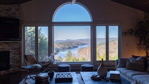 Een woonkamer met een groot rondboogvenster met uitzicht op een rivier en heuvels. Er is een open haard, een bank en een salontafel met boeken en decoratieve objecten.