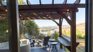 Een houten terras met buitenmeubilair, waaronder stoelen en een bankje, met uitzicht op een meer en heuvels.