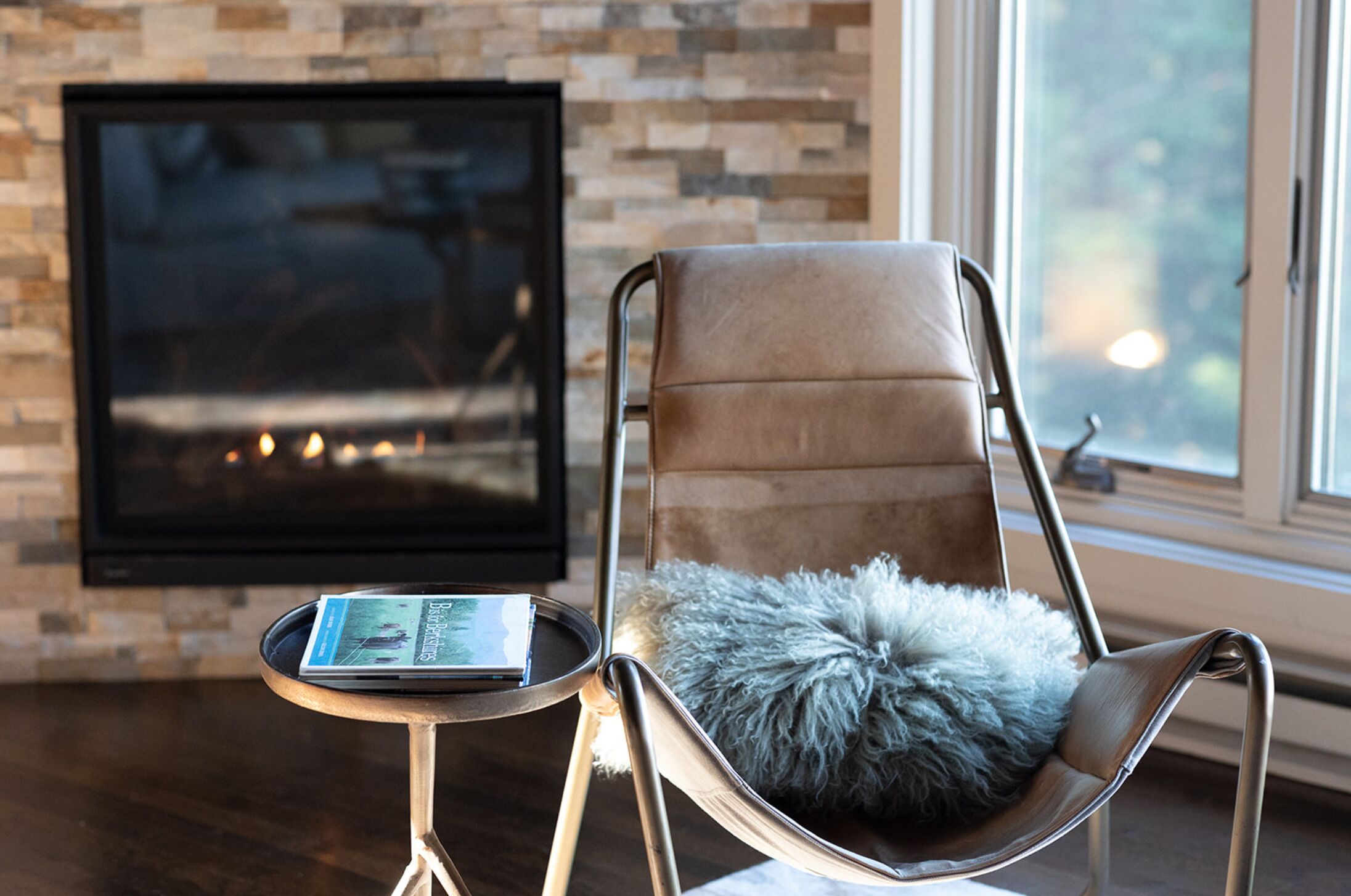 A modern living room with a fireplace, a leather chair with a fluffy pillow, and a small table with a magazine.