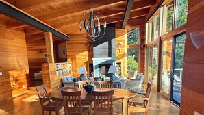 Interior de una cabaña de madera con comedor, chimenea y ventanas amplias.