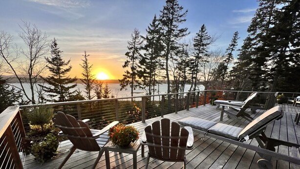 Una terraza con sillas tipo Adirondack y una mesa, con vista a un cuerpo de agua y árboles.