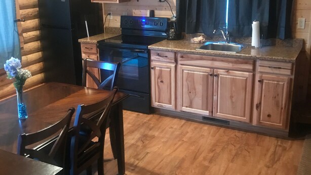 A log cabin kitchen with wooden cabinets, a microwave, a stove, and a dining area with a table and chairs. - Cabin #8 (Garden City)