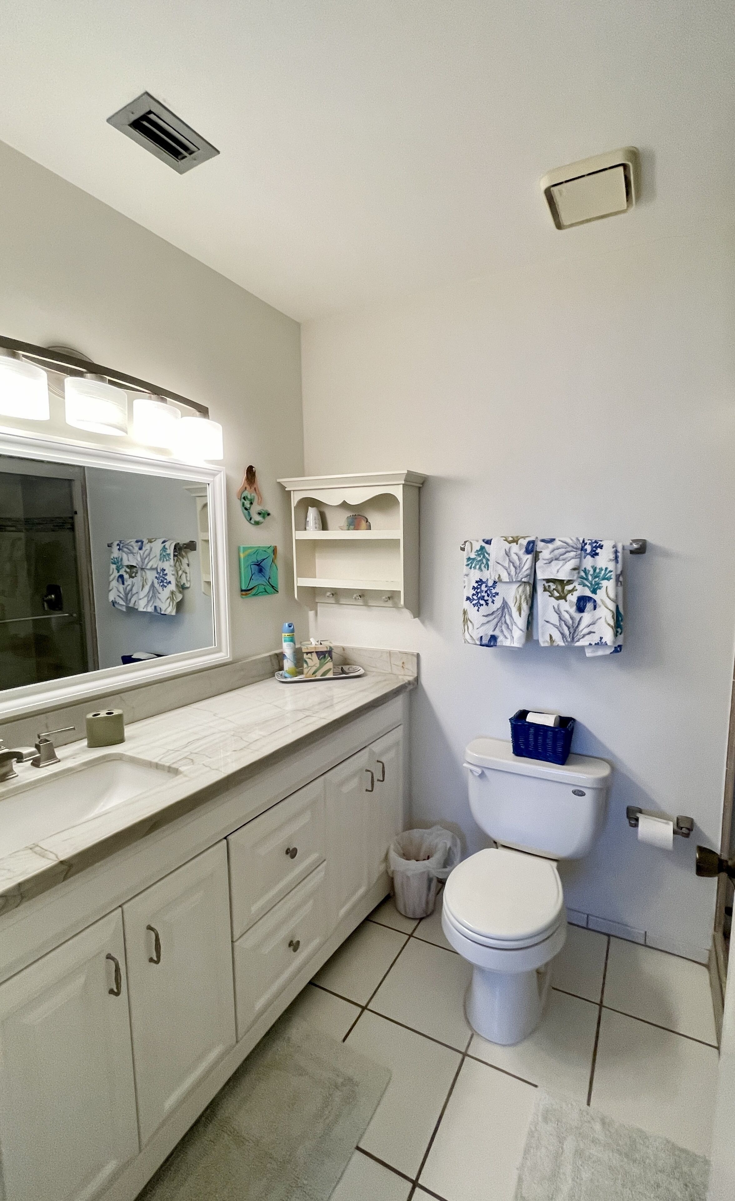 Une salle de bain avec un lavabo blanc, un miroir et des toilettes. On y trouve deux porte-serviettes avec des serviettes, une poubelle et une étagère au-dessus des toilettes.