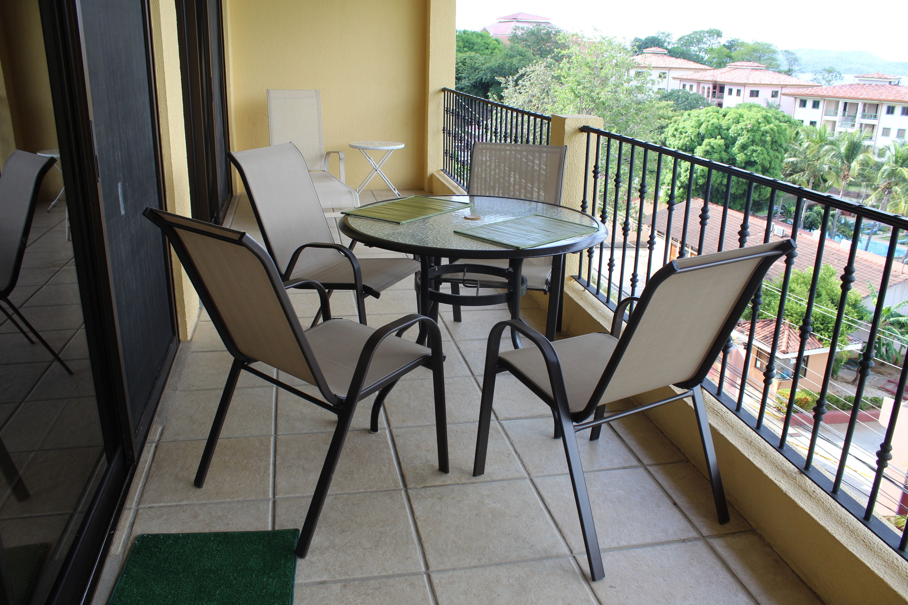 A balcony with a round table and four chairs, a small folding table, and a green doormat.