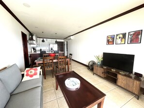 A living room with a sofa, dining table, chairs, a TV, and a kitchen in the background.