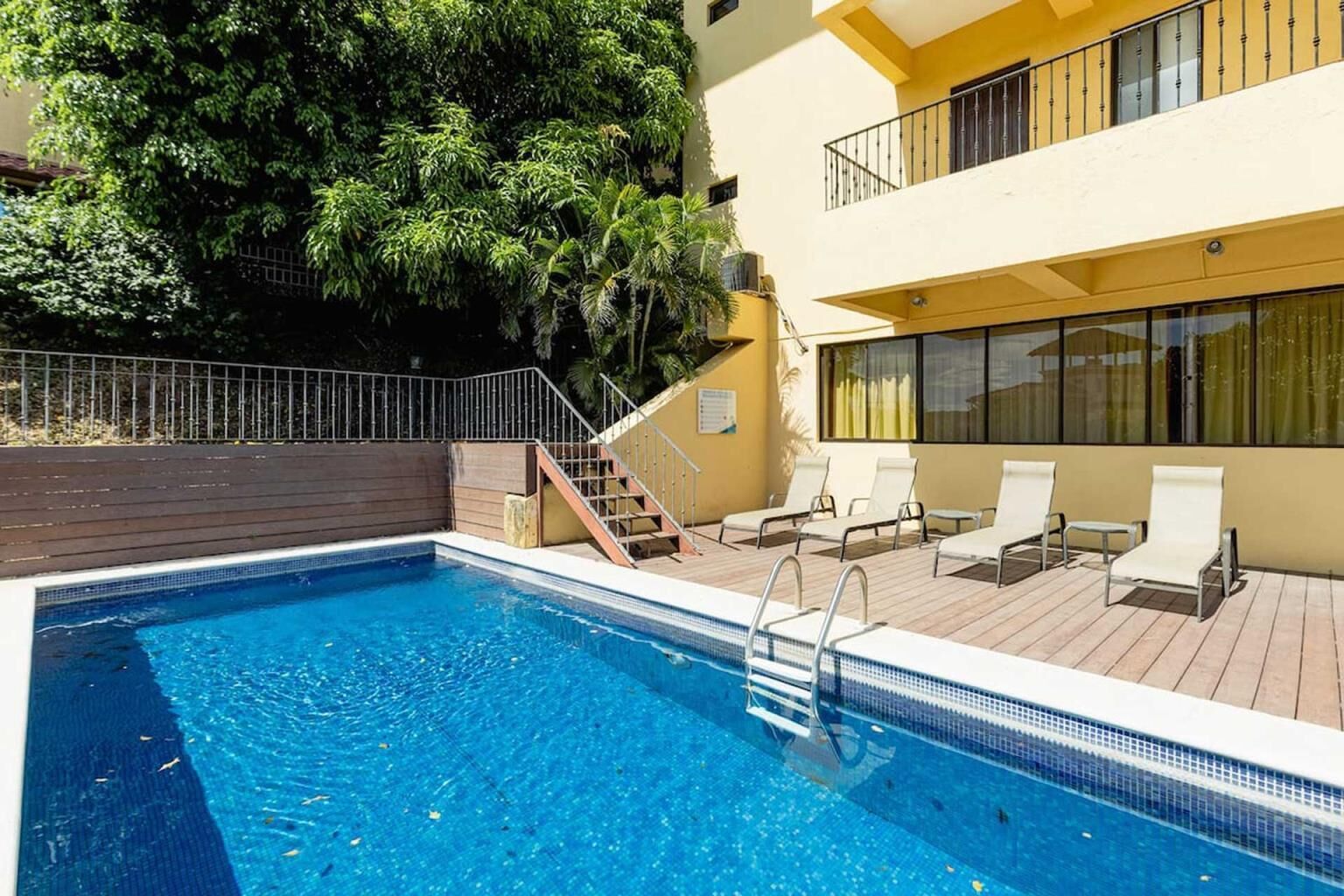 Une piscine entourée d’une terrasse en bois, de chaises longues et d’un escalier menant à un balcon.