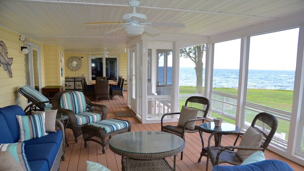 A covered outdoor seating area with wicker furniture, a glass table, and a view of the ocean. - Sunset Lodge on the Bay (Rock Hall)