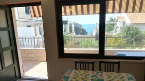 Une pièce avec une table recouverte d’une nappe à motifs floraux, deux fenêtres dotées de stores à rayures et une vue sur la mer.