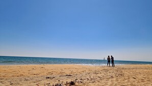 Una playa con dos personas de pie sobre la arena, un velero a lo lejos y un cielo azul despejado.