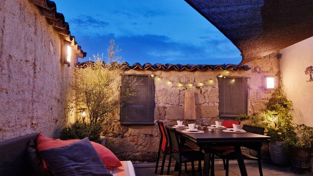 Un patio al aire libre con un muro de piedra, una mesa de madera dispuesta para comer, una zona de estar con cojines e iluminación con luces colgantes.