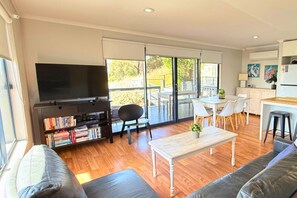 A modern living room with a flat-screen TV, bookshelves, a sofa, a coffee table, and a sliding glass door leading to an outdoor patio. - Lake & Reserve Front Bird Haven - 3BR (Encounter Bay)