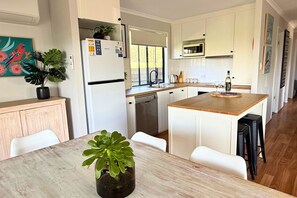 A modern kitchen with white cabinetry, a central island, and a dining area with a table and chairs. - Lake & Reserve Front Bird Haven - 3BR (Encounter Bay)
