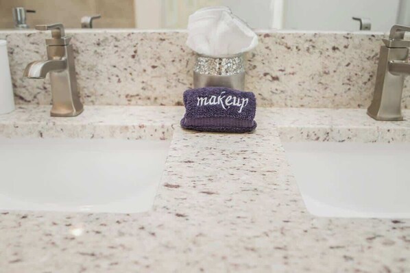 Une salle de bain avec un comptoir en marbre, deux lavabos et un porte-serviettes sur lequel sont posés une serviette blanche et une serviette violette avec le mot « maquillage ».