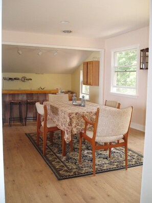Un comedor con una mesa dispuesta para cuatro, sillas de madera, un tapete estampado y una cocina al fondo.