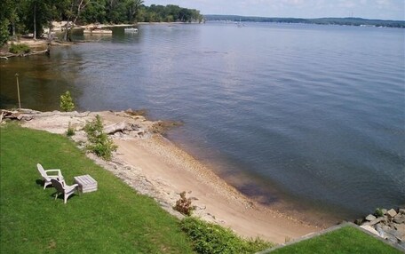A lakeside view with a grassy area, a sandy beach, and a row of white chairs.