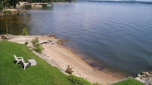 A lakeside view with a grassy area, a sandy beach, and a row of white chairs. - 5-bedroom vacation home 5 minutes from historic Charlestown (Perryville)