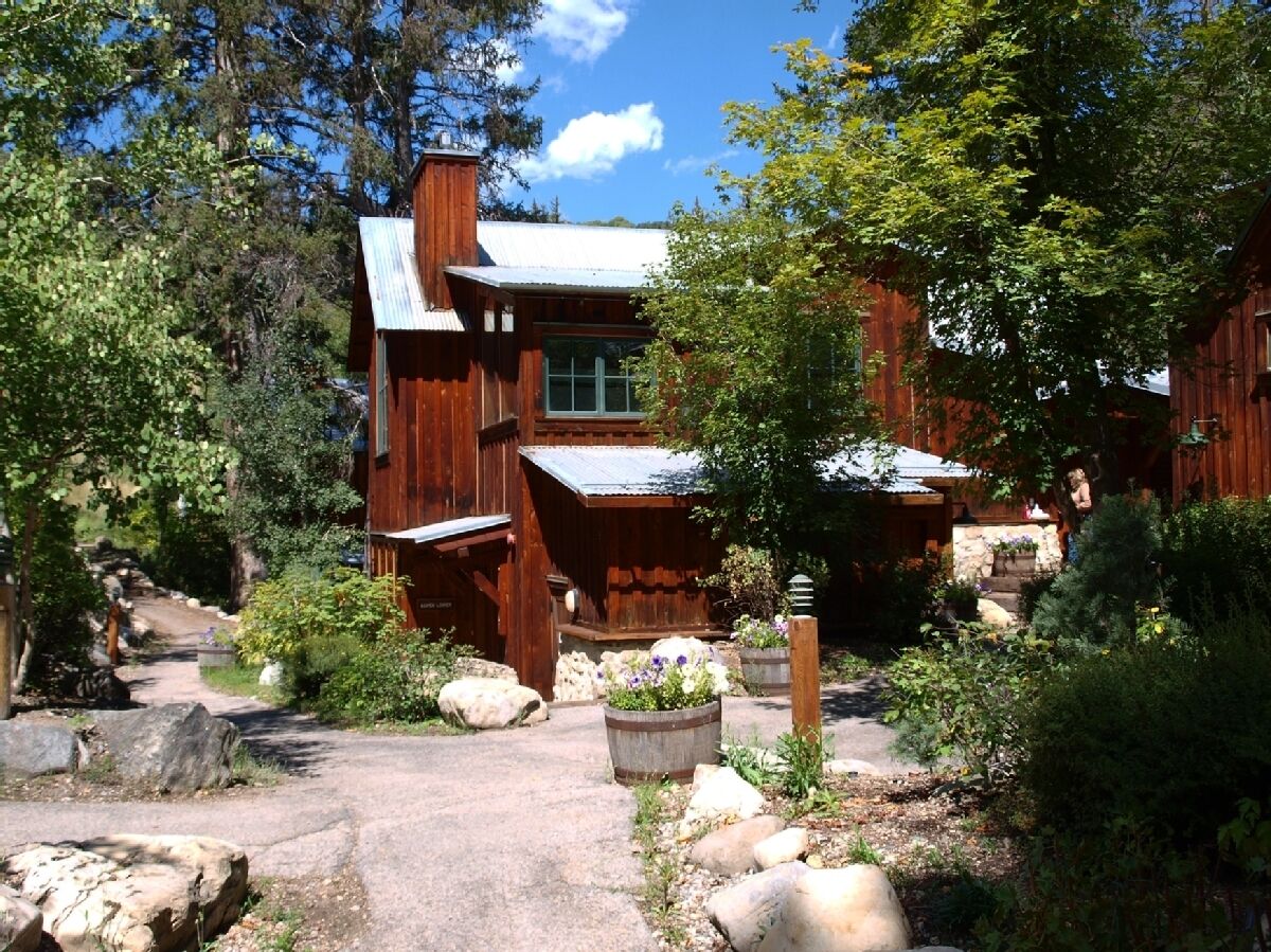 A rustic wooden cabin with a stone pathway leading to the entrance, surrounded by lush greenery and large rocks.