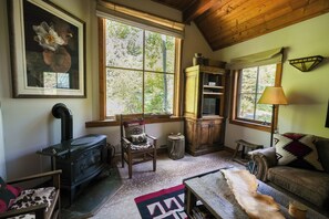 A cozy room with a wood-burning stove, a chair, a sofa, a rug, and a window with a view of trees. - Luxury Cottage at Sundance Utah-Book Fall Leaf Stay-Walk to Lift and Restaurants (Sundance)