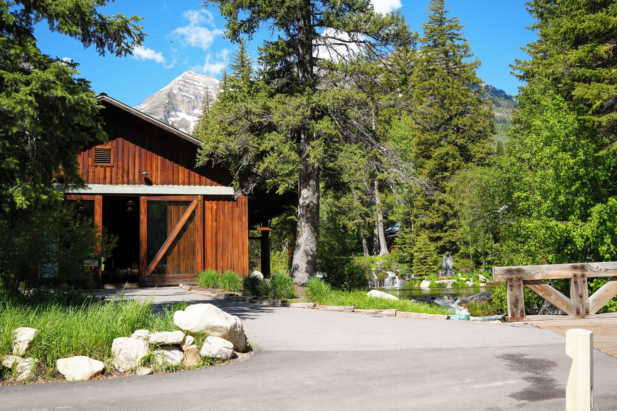 A rustic wooden building with a large entrance, surrounded by lush greenery and a mountain backdrop.