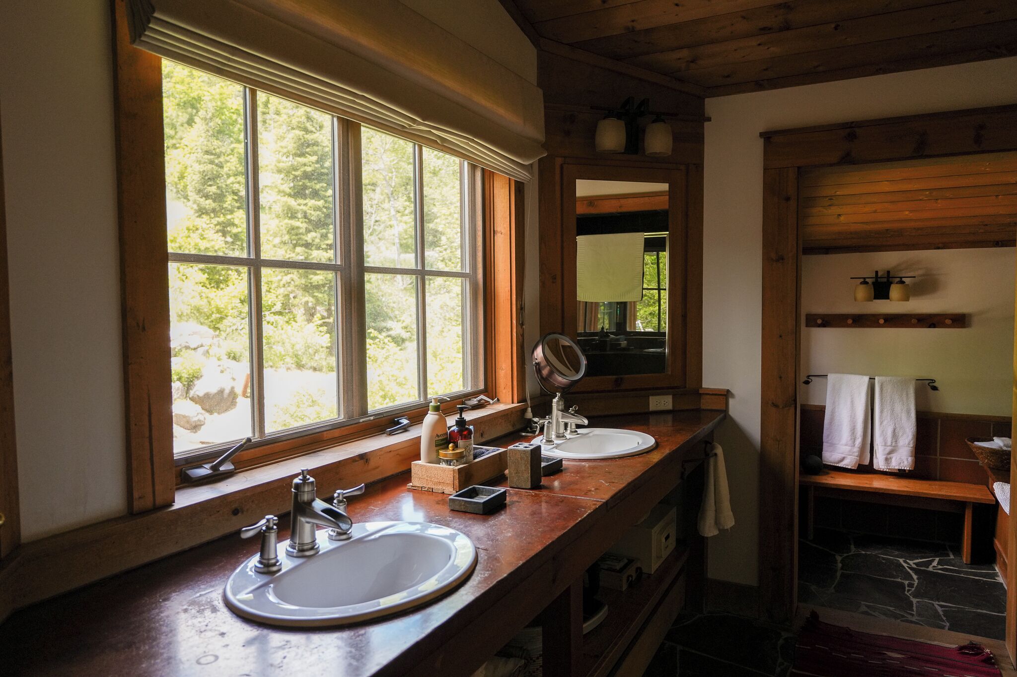 Un baño con dos lavamanos, una ventana con vistas a árboles, un espejo y un perchero para toallas.
