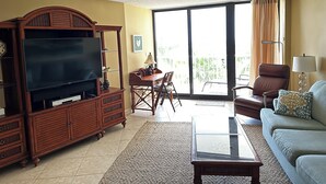 A living room with a TV, wooden furniture, a glass coffee table, a sofa, and a balcony with curtains. - Beachfront 2BR in South Seas T3-508 with Updated Kitchen! (Marco Island)