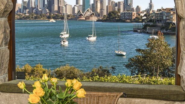 Uma mesa de vidro com um prato de comida, uma cadeira de vime e um vaso de flores, com vista para um porto com barcos e o horizonte da cidade.