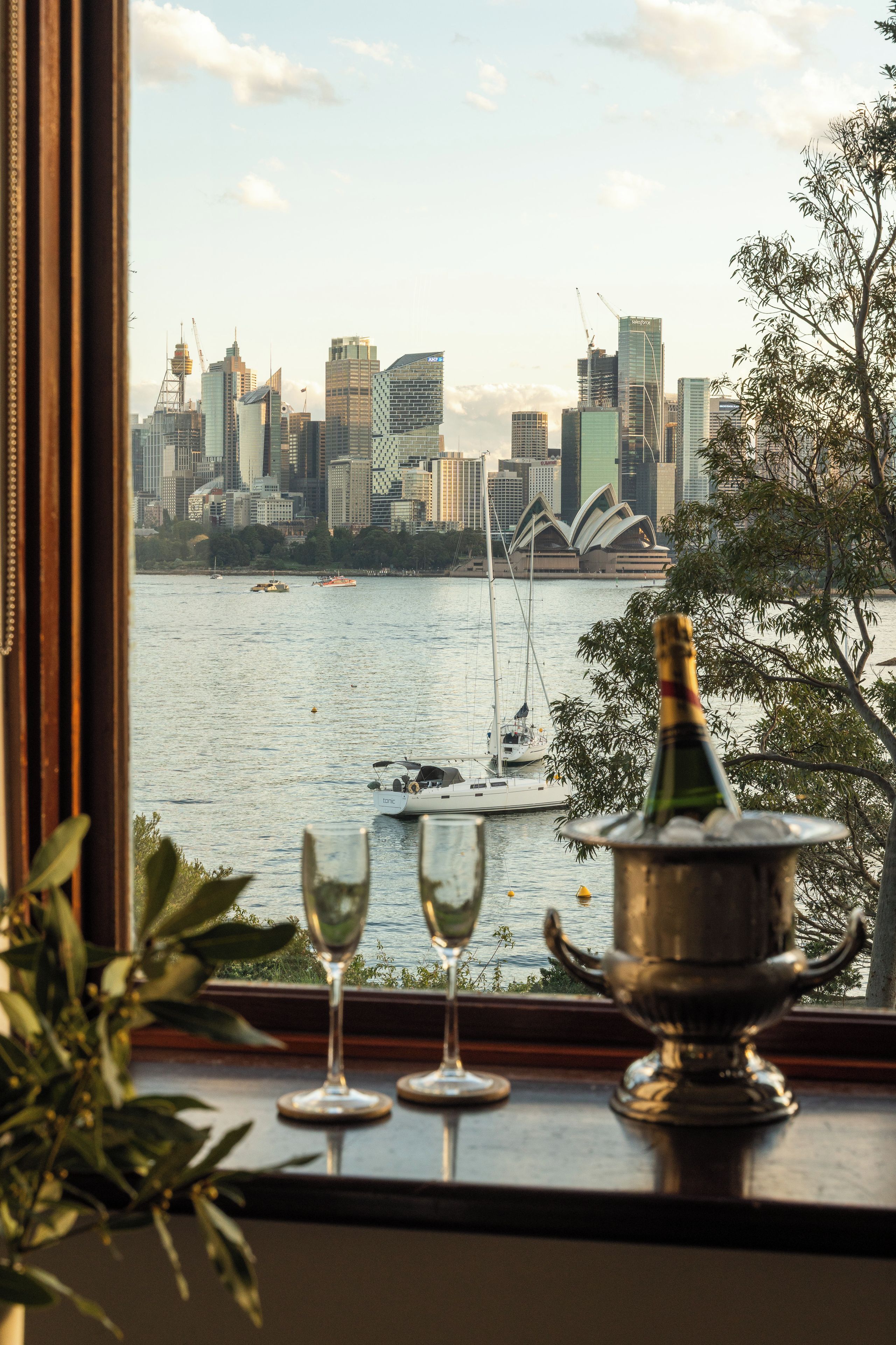 A view of a city skyline with a body of water in the foreground, a bottle of champagne, and two wine glasses on a windowsill.