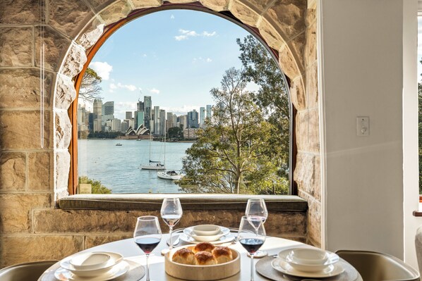 A table with wine glasses and bread, overlooking a city skyline and harbor through an arched window.