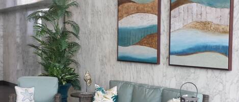 Un vestíbulo con pared de mármol, dos cuadros de paisajes enmarcados, muebles de mimbre, una planta en maceta y una mesita con adornos.