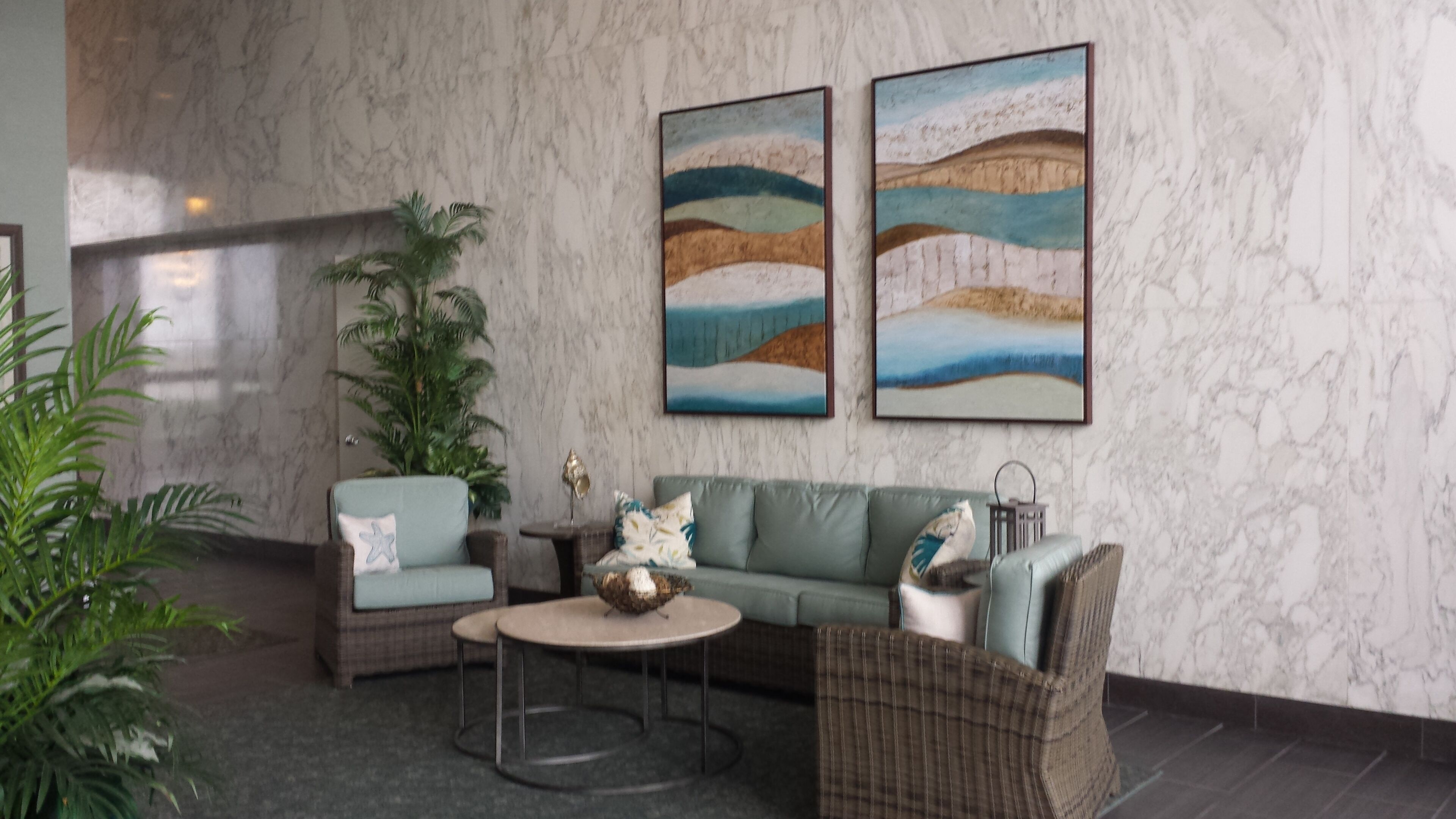 A lobby with a marble wall, a green sofa, a wicker chair, a round table, and two paintings.