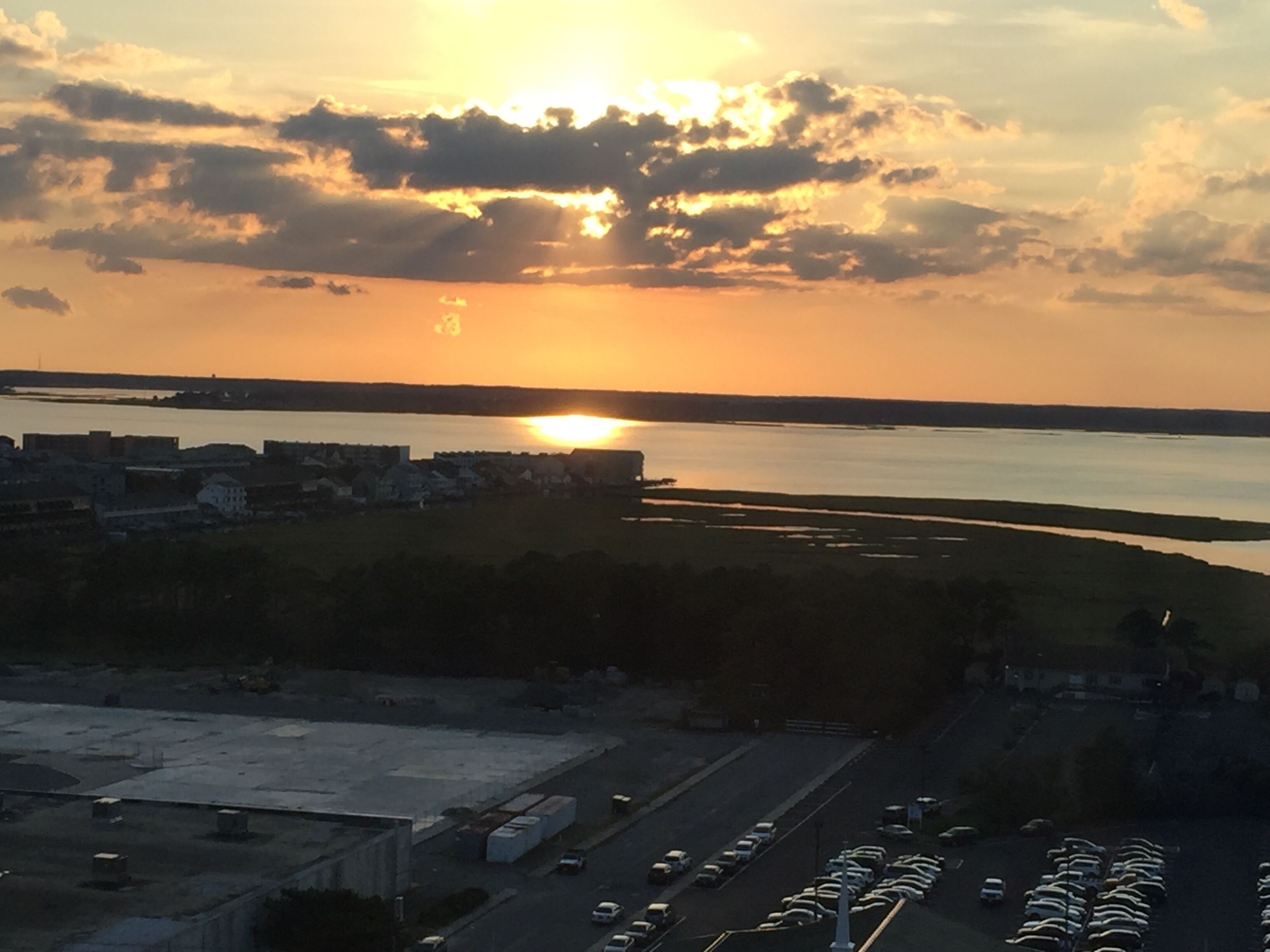 Vista aerea di una città costiera al tramonto, con un’ampia distesa d'acqua, edifici industriali e un parcheggio.