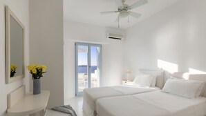 Ein modernes Schlafzimmer mit zwei Betten, einem Deckenventilator, einer Klimaanlage, einem kleinen Tisch mit Lampe und Meerblick durch eine Glastür.