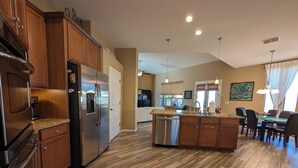 A spacious kitchen with wooden cabinets, a stainless steel refrigerator, a dishwasher, and a dining area with a glass island and chairs. - Casa Bella-Luxury home on corner lot, bring your fur baby. (Oro Valley)