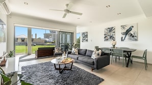 A modern living room with a grey sofa, a dining table, and chairs. There is a large window with a view of the outside. - Pilotage 3 - Lighthouse Views & Coastal Living in the Heart of Yamba - Holiday Yamba (YAMBA)