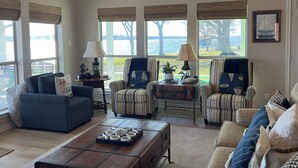 Ein geräumiges Wohnzimmer mit einem blauen Sofa, gestreiften Sesseln und einem Couchtisch. Große Fenster bieten Blick auf einen See und Bäume.