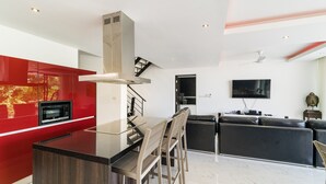 A modern kitchen with a central island, a dining area with chairs, a black sofa, and a TV mounted on the wall. - 3 Bedroom Sea View Villa Mattana 2 SDV162 - Chaweng Noi - By Samui Dream Villas (Koh Samui)