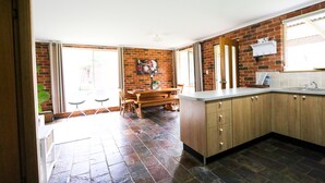 A kitchen with a brick wall, wooden cabinets, a dining area with a bench, and a large window. - Mogendoura Farm 3 COTTAGES MAX 12 ADULTS but 18 people including children (Moruya)