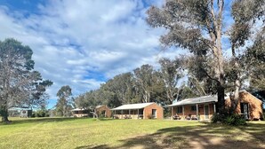 A rural setting with a large open grassy area, a dirt path, and several brick buildings with attached porches, surrounded by trees. - Mogendoura Farm 3 COTTAGES MAX 12 ADULTS but 18 people including children (Moruya)