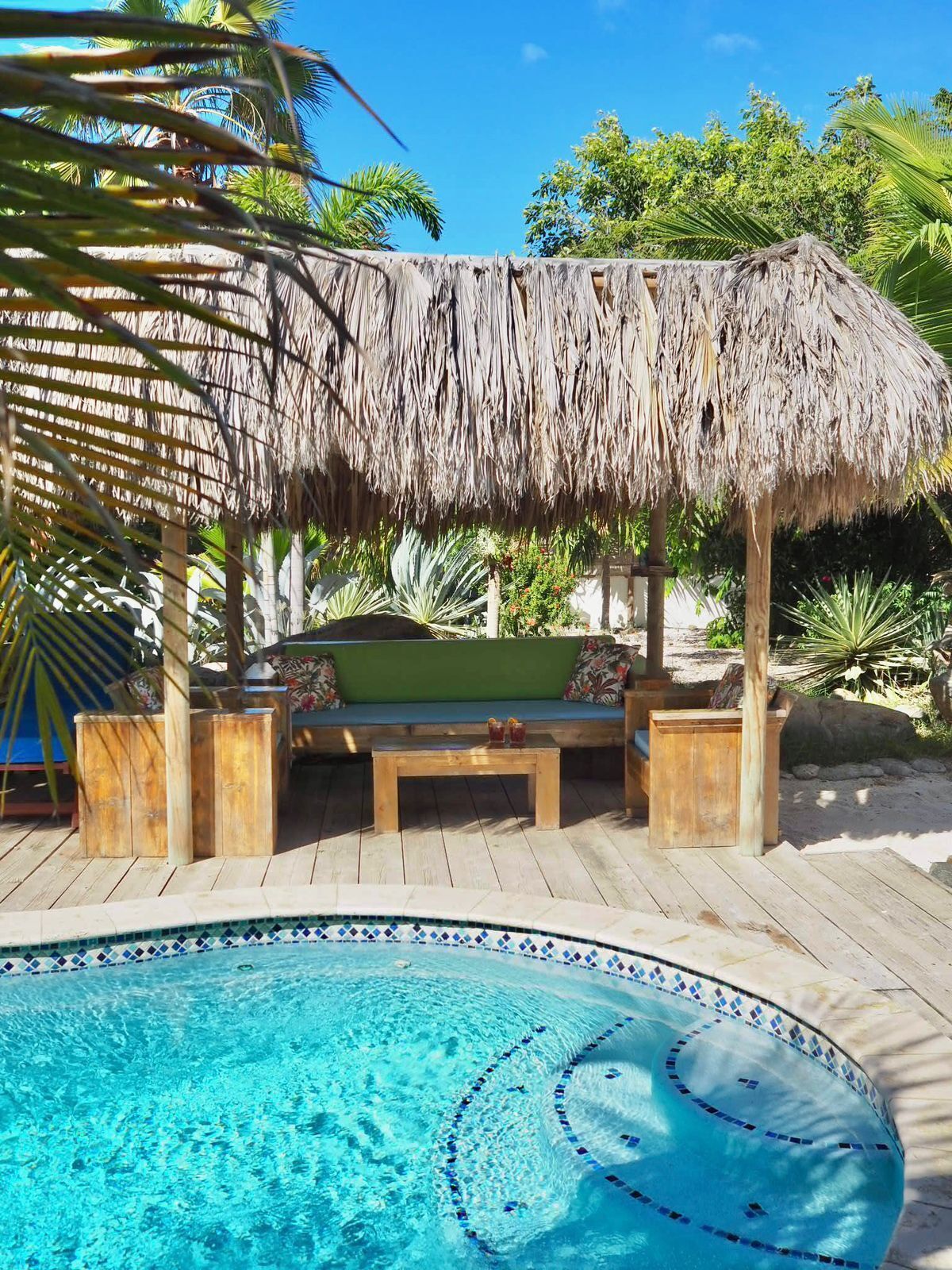 Un espace aménagé au bord de la piscine, doté d’un coin salon avec un toit de chaume, d’une table en bois et d’une piscine aux carreaux de mosaïque bleus.