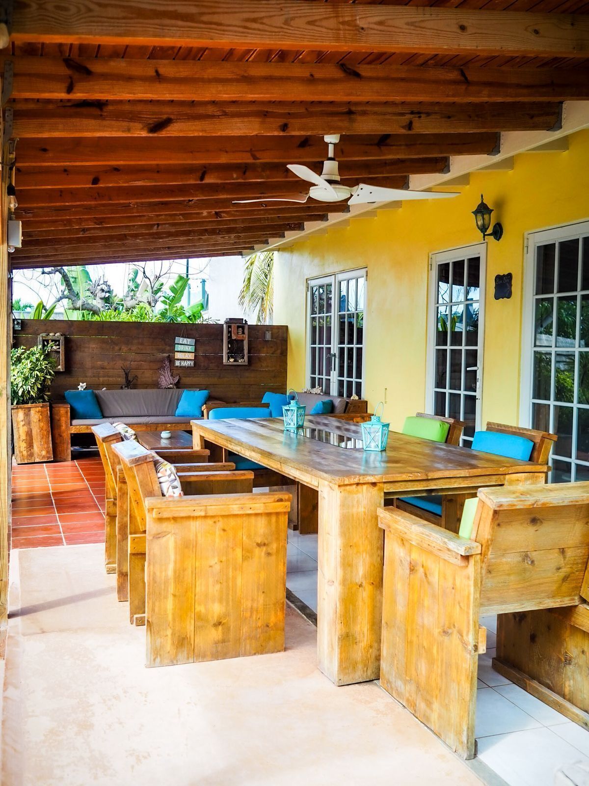 An outdoor patio with wooden furniture, a ceiling fan, and a yellow wall.
