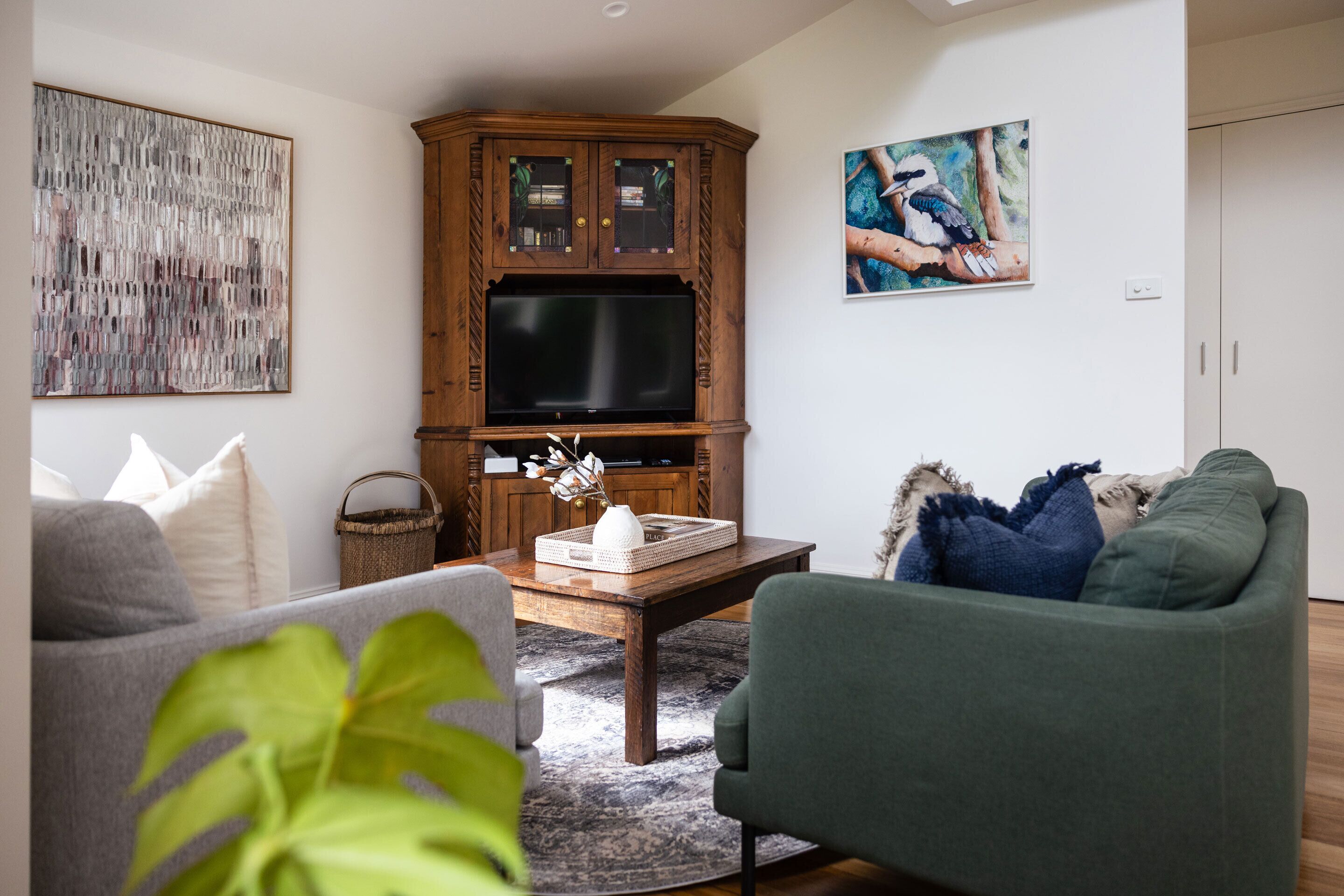 A living room with a wooden cabinet, a flat-screen TV, a grey sofa, a green armchair, and a coffee table with decorative items.