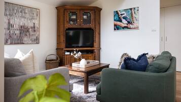 A living room with a wooden cabinet, a flat-screen TV, a grey sofa, a green armchair, and a coffee table with decorative items.