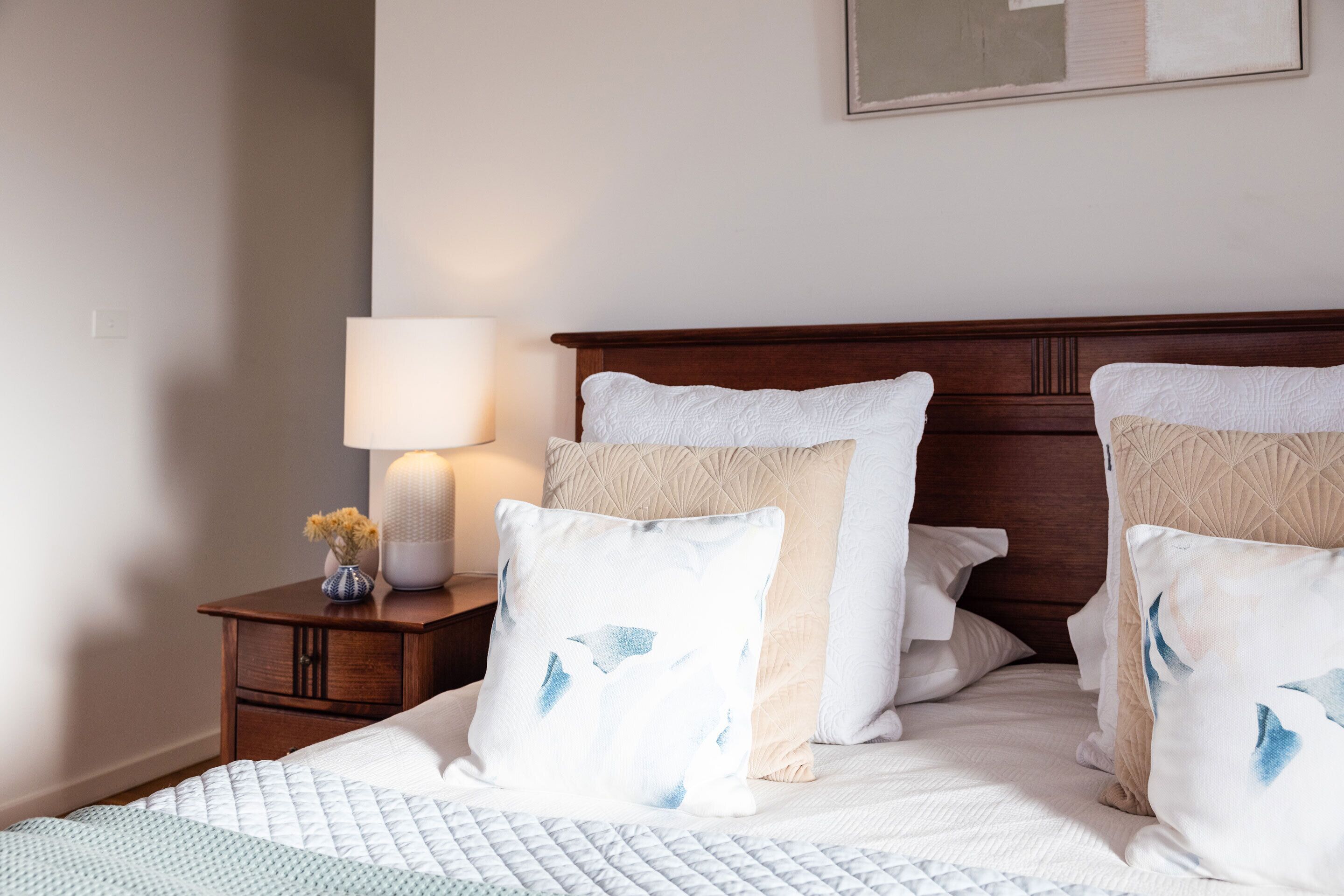 A neatly made bed with white and blue patterned pillows, a wooden headboard, and a bedside table with a lamp and a vase of flowers.