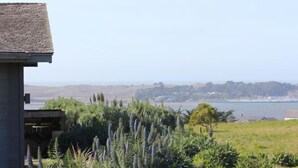 Eine Küstenlandschaft mit einem Gebäude mit geneigtem Dach und Blick auf das Meer und die dahinterliegenden Hügel.