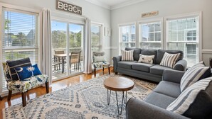 A spacious living room with a gray sofa, a patterned rug, and a view of the outdoors through large windows. - Water's Edge 103 - Bright 3BR/3BA Townhome - Pool - Fishing Dock (Folly Beach)