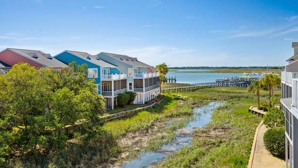 A coastal residential area with multi-story buildings, a waterfront view, and a dock extending into the water. - Water's Edge 103 - Bright 3BR/3BA Townhome - Pool - Fishing Dock (Folly Beach)