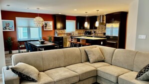 A modern living room with a beige sectional sofa, a dining area with a table and chairs, and a kitchen with dark cabinets and stainless steel appliances.